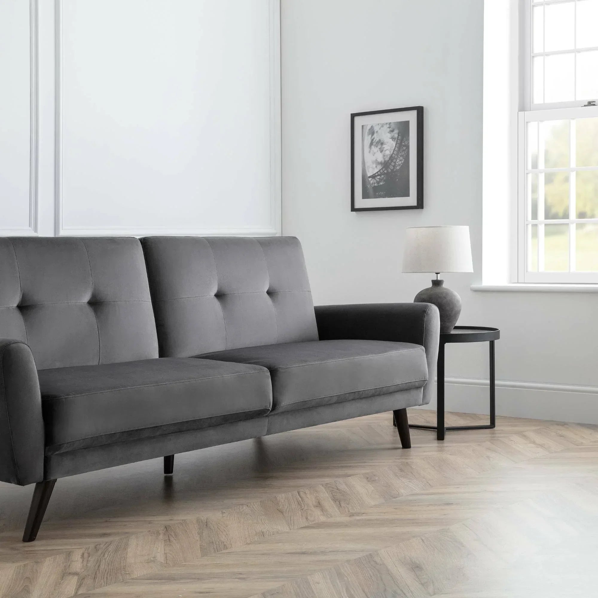 Grey velvet sofabed with tufted seat, wooden legs, next to lamp table in bright living room