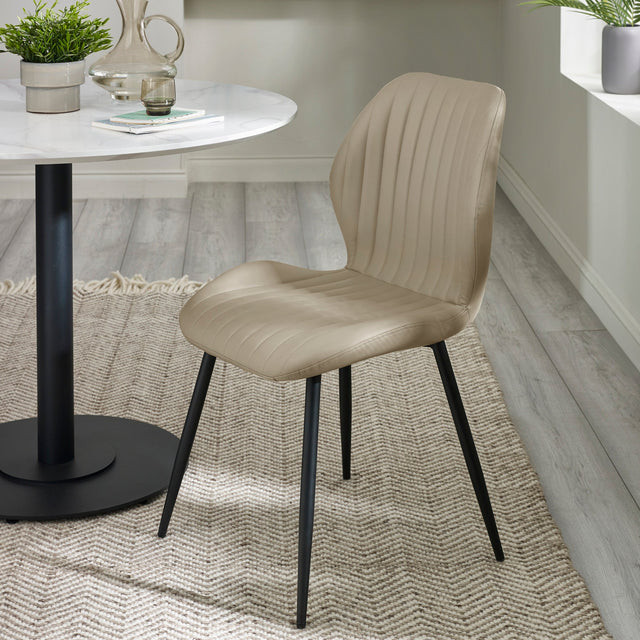 Beige upholstered dining chair with vertical stitching and black tapered metal legs beside round white table