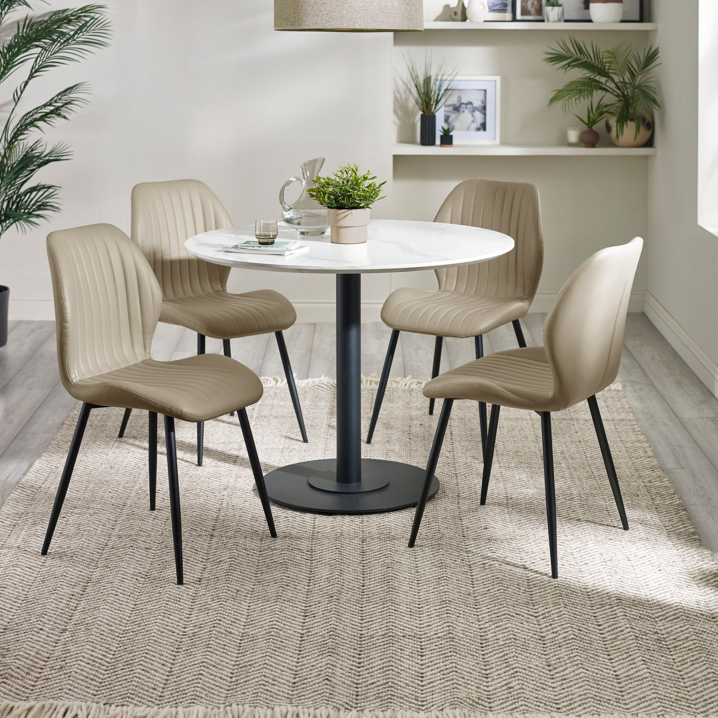 Four beige upholstered dining chairs with black legs around a round white table in a room with light wood floors and shelves.