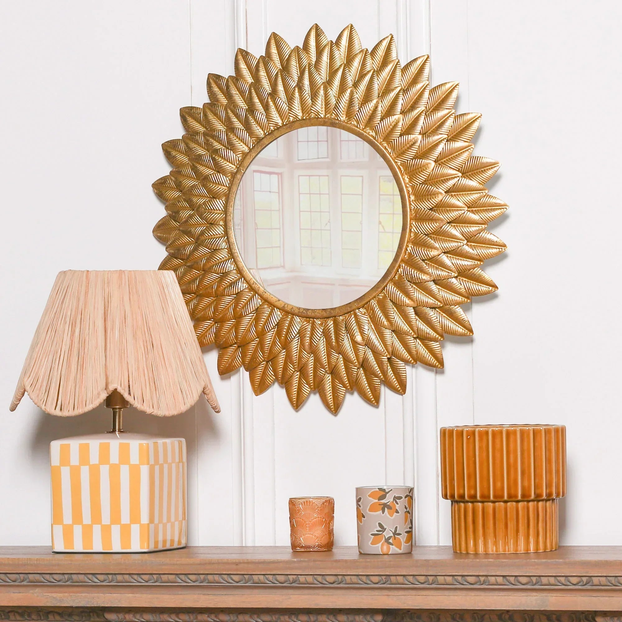 Gold leaf sunburst wall mirror above mantel with decorative lamp and orange vases