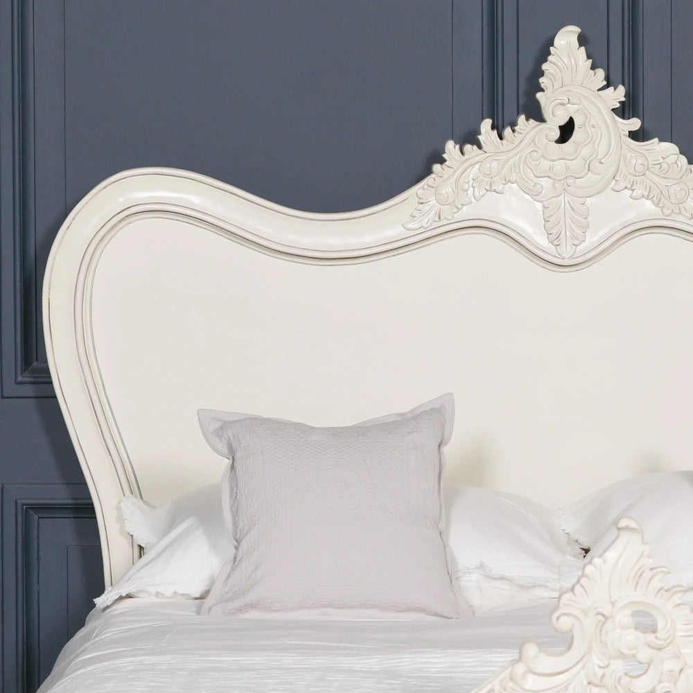 Cream French style bed with ornate carved headboard and neutral bedding against dark wall