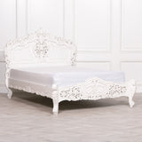 A white ornate carved wooden bed frame with a matching headboard and footboard, shown with a white mattress on a light gray wood floor against a white paneled wall.