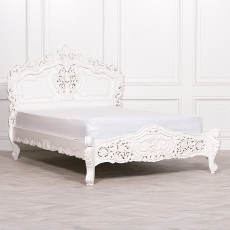 A white ornate carved wooden bed frame with a matching headboard and footboard, shown with a white mattress on a light gray wood floor against a white paneled wall.