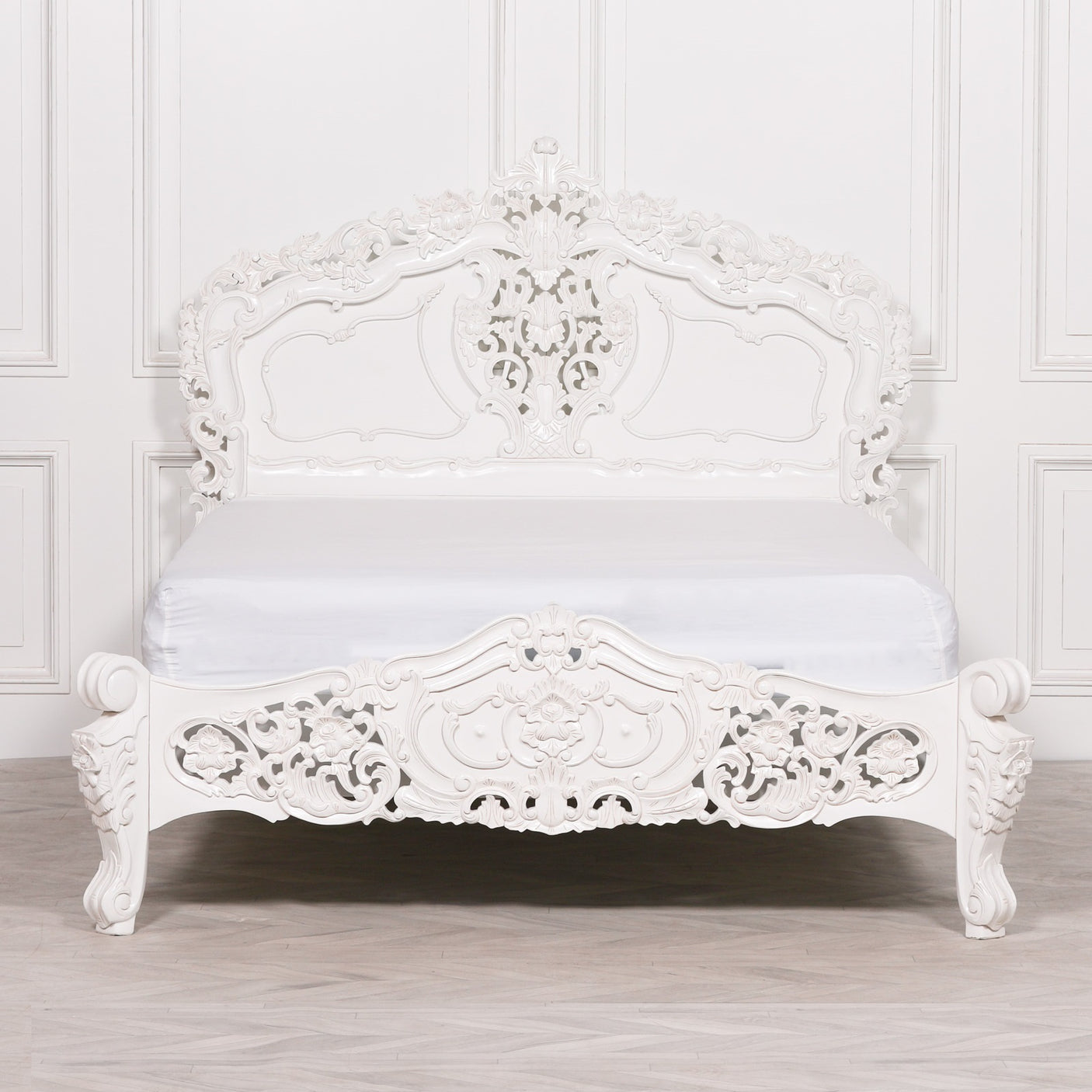 An ornate white carved wooden bed frame with a matching white mattress, set against a white paneled wall and light wood floor.