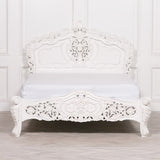An ornate white carved wooden bed frame with a matching white mattress, set against a white paneled wall and light wood floor.
