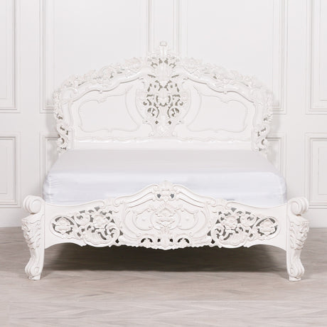 An ornate white carved wooden bed frame with a matching white mattress, set against a white paneled wall and light wood floor.