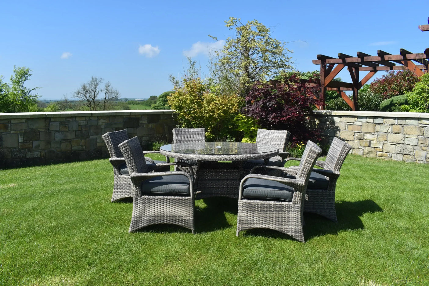Grey rattan dining set with six cushioned chairs and round glass table on a garden lawn