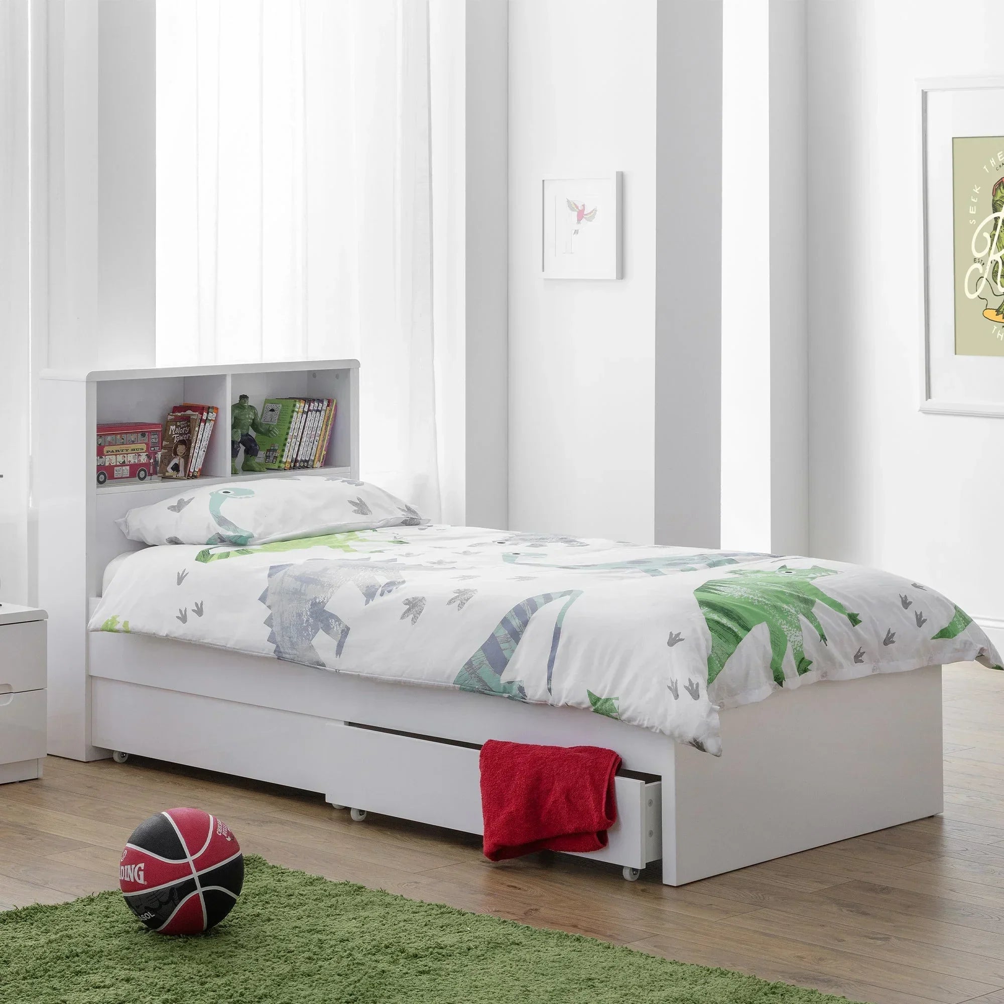 White bookcase bed with underbed storage drawer, dinosaur bedding, and books on headboard