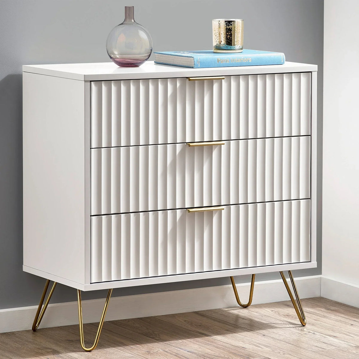 White three drawer chest with ribbed fronts and gold handles on gold hairpin legs