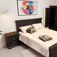 A dark wood bed frame with white bedding and two patterned pillows, styled in a bedroom setting with a matching nightstand, a wall lamp, and a colorful abstract painting on the wall.
