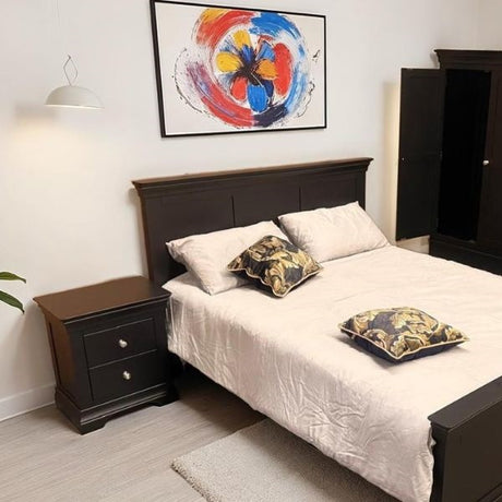 A dark wood bed frame with white bedding and two patterned pillows, styled in a bedroom setting with a matching nightstand, a wall lamp, and a colorful abstract painting on the wall.