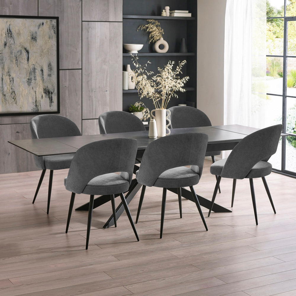 Extended black dining table with six grey velvet chairs in bright room with floor-to-ceiling windows and decor.