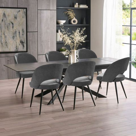 Extended black dining table with six grey velvet chairs in bright room with floor-to-ceiling windows and decor.