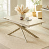 Light beige extending dining table with crossed metal legs in cream, set in bright room on textured beige rug
