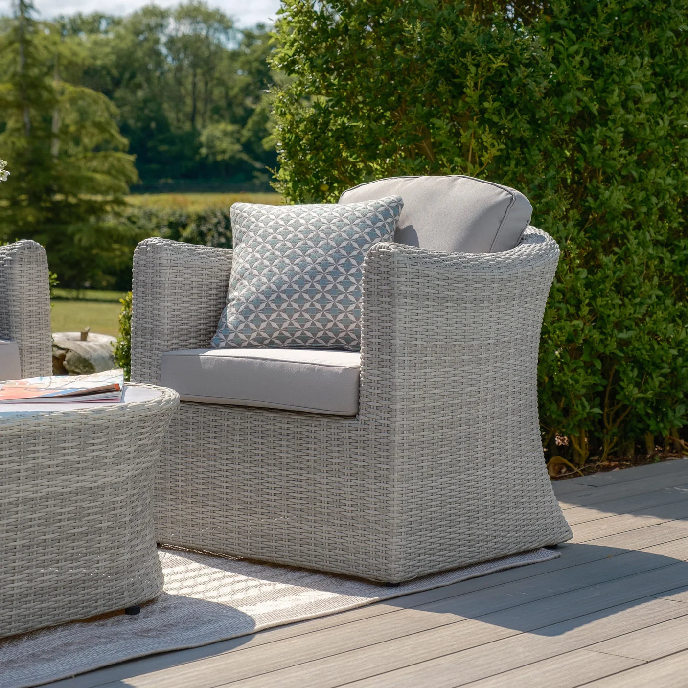 Light grey rattan garden armchair with seat cushion and patterned pillow on decking