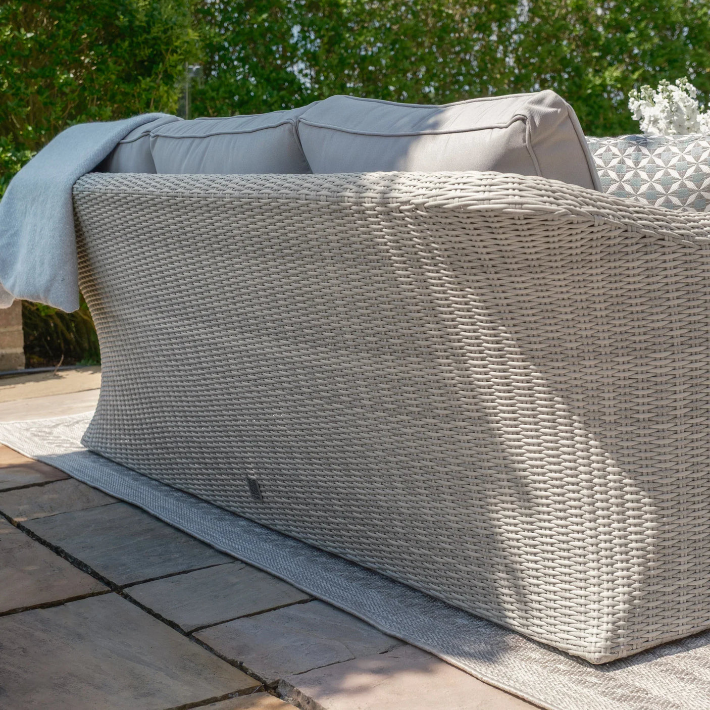 Light grey rattan outdoor sofa with cushions on stone patio facing garden greenery