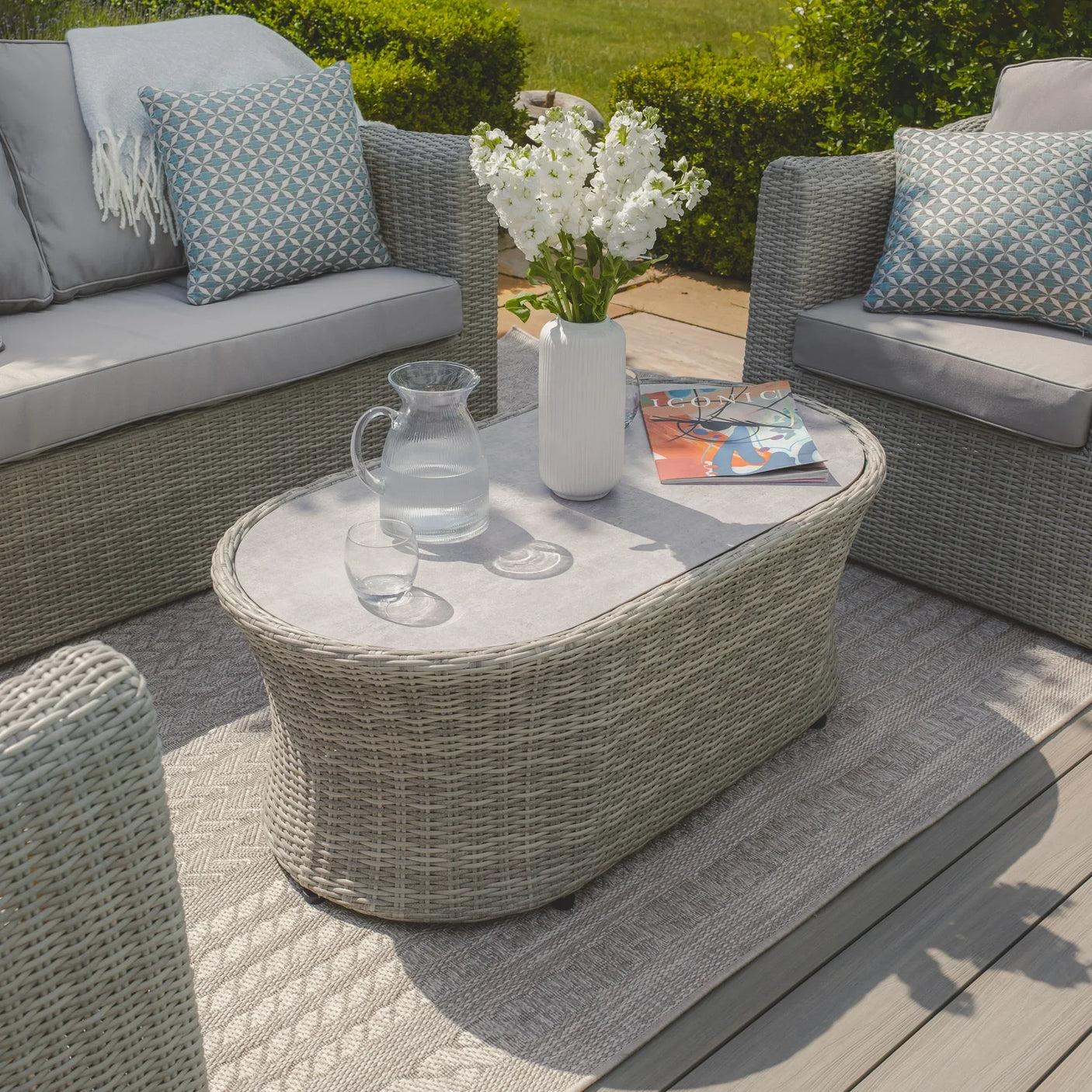 Grey rattan garden coffee table with glass top, vase, and pitcher, surrounded by outdoor seating