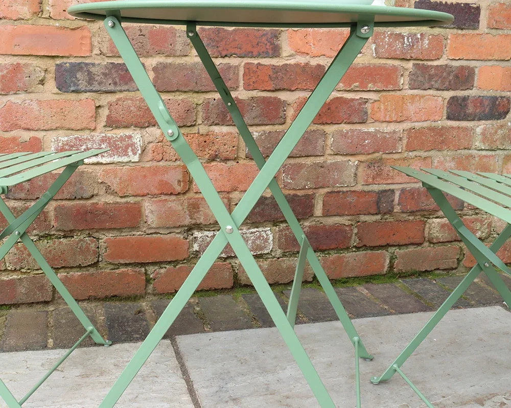 Olive green metal bistro table and two chairs set against a brick wall outdoors