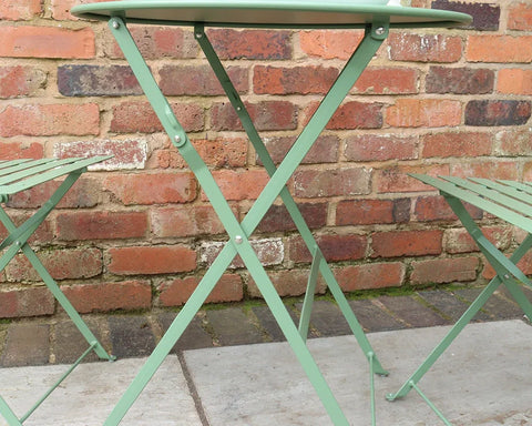 Olive green metal bistro table and two chairs set against a brick wall outdoors