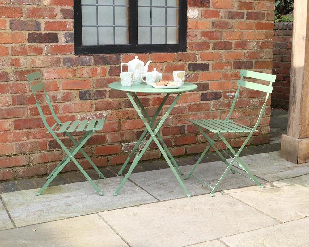 Olive green metal bistro set with round folding table and two chairs on patio by brick wall