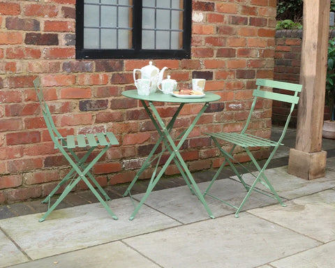 Olive green metal bistro set with round table and two chairs on patio against brick wall
