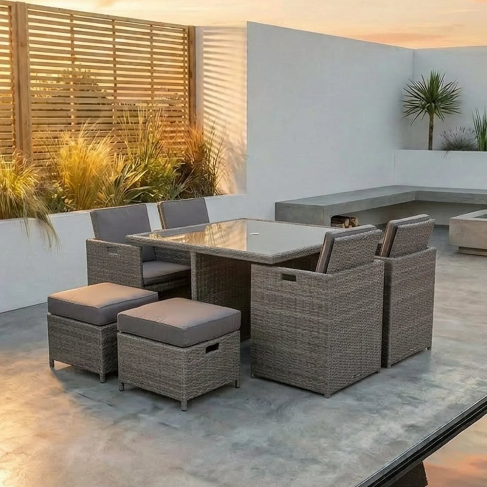 Rattan outdoor dining set with glass table, cushioned chairs and stools on a patio at sunset