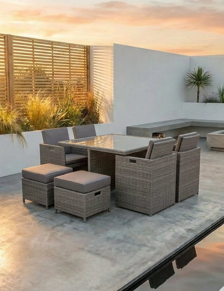 Rattan outdoor dining set with glass table, cushioned chairs and stools on a patio at sunset