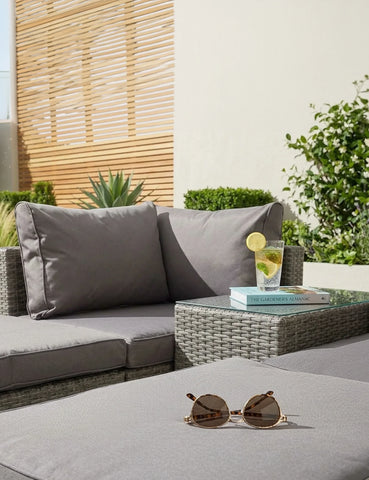 Grey rattan garden sofa set with cushions, side table, sunglasses, and glass of drink outdoors