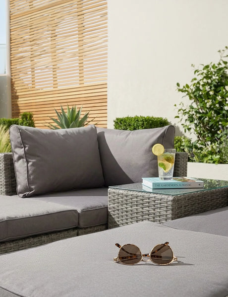 Grey rattan garden sofa set with cushions, side table, sunglasses, and glass of drink outdoors