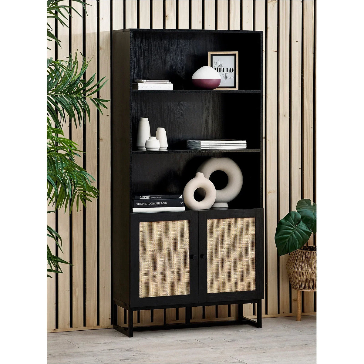 Black tall bookcase with rattan doors, open shelves, decor accents against wood panel wall