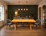 A rustic wooden dining table with four gray upholstered chairs in a styled dining room with a dark green paneled wall, pendant lights, and a sideboard.