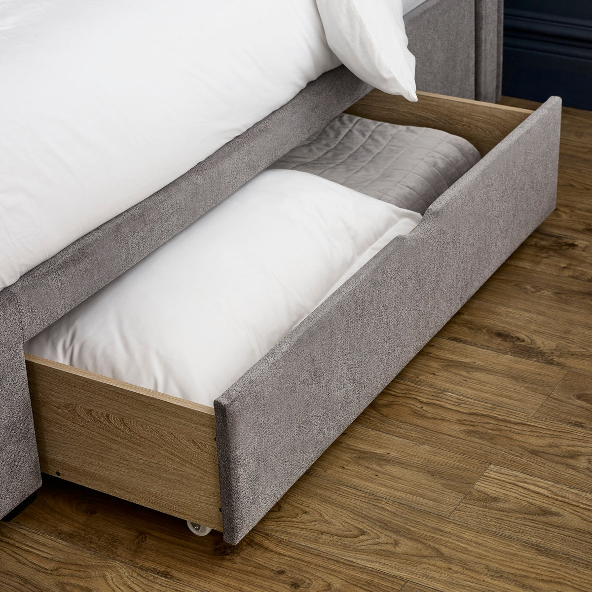 Grey fabric storage drawer open under bed with white bedding inside on wooden floor