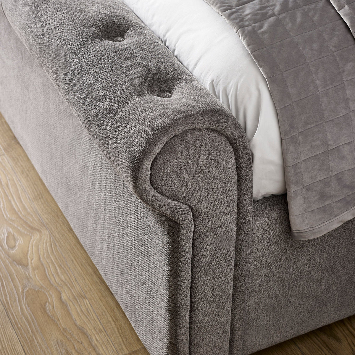 Close-up of curled armrest on grey fabric button-tufted storage bed on wooden floor