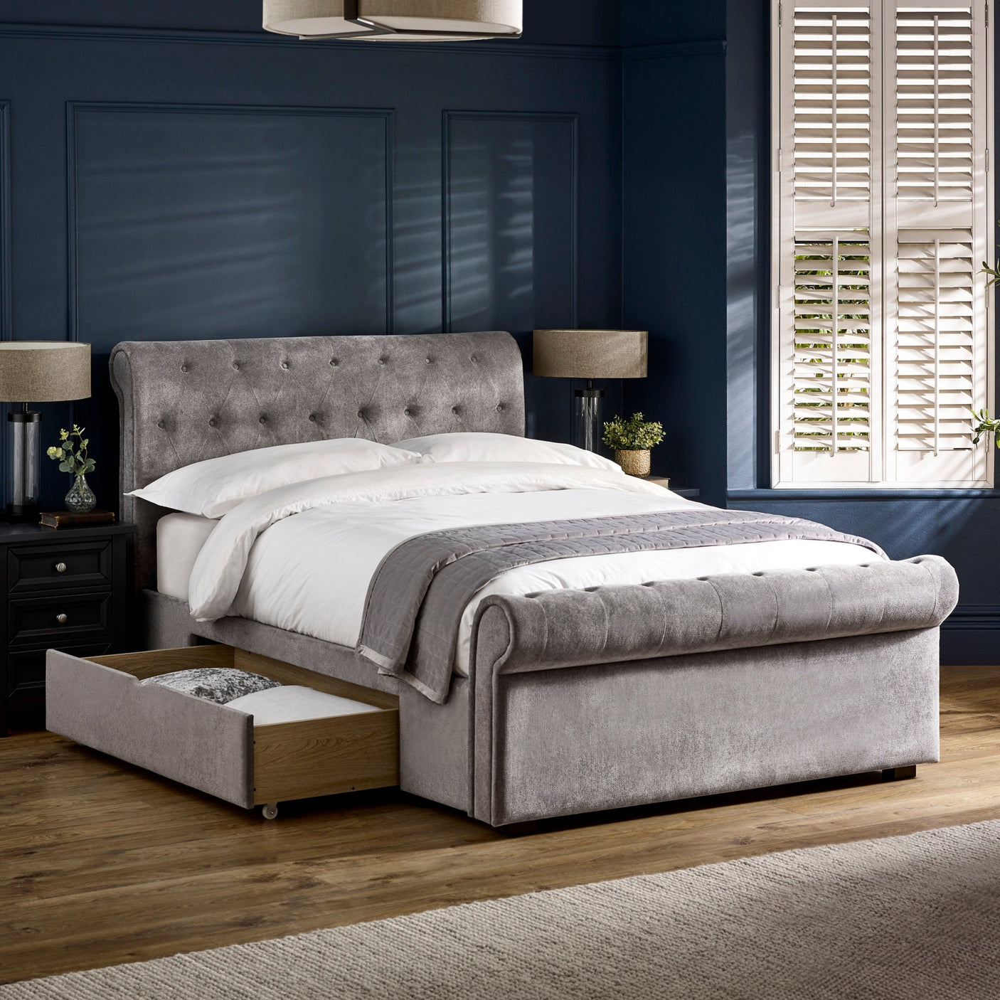 Same grey fabric double bed with left side drawer opened, showing storage space in a dark blue room.