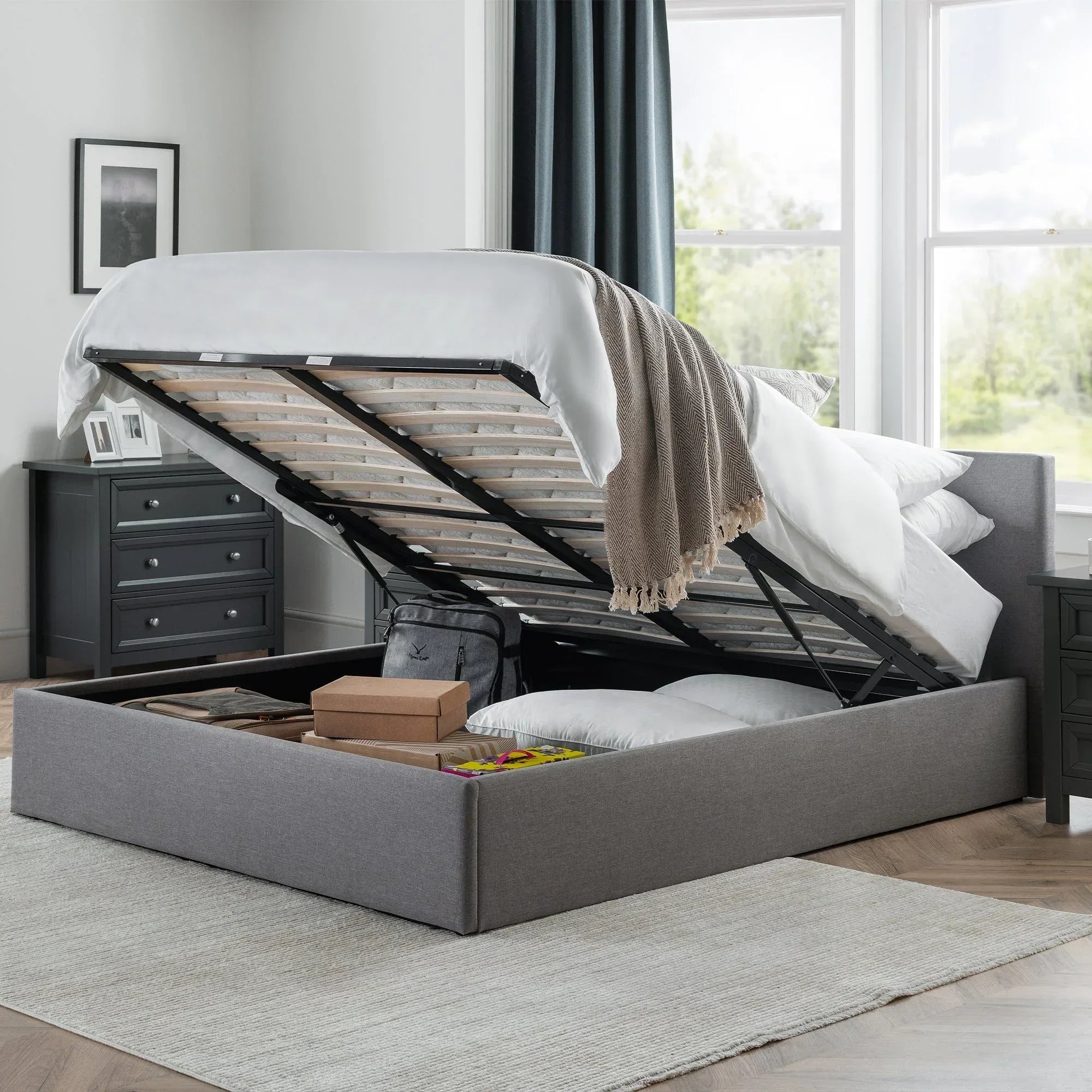 Grey lift-up storage bed with open base showing storage boxes and pillows in bedroom
