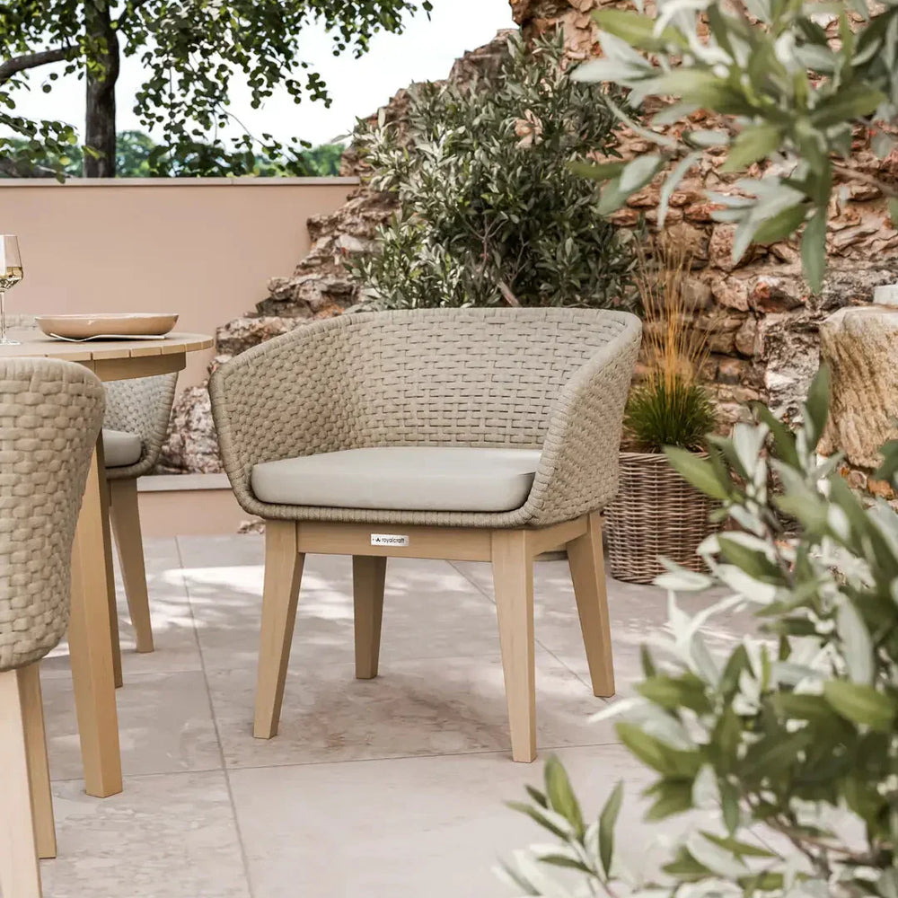 Beige outdoor woven dining chair with cushion on patio, surrounded by greenery and stone wall