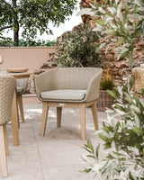 Beige outdoor woven dining chair with cushion on patio, surrounded by greenery and stone wall