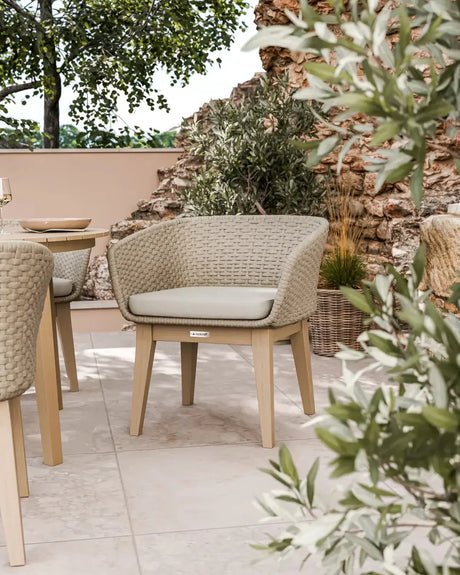 Beige outdoor woven dining chair with cushion on patio, surrounded by greenery and stone wall