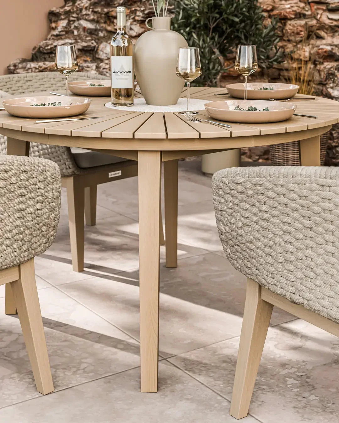 Round wood outdoor dining table with rope chairs set for four on patio tiles