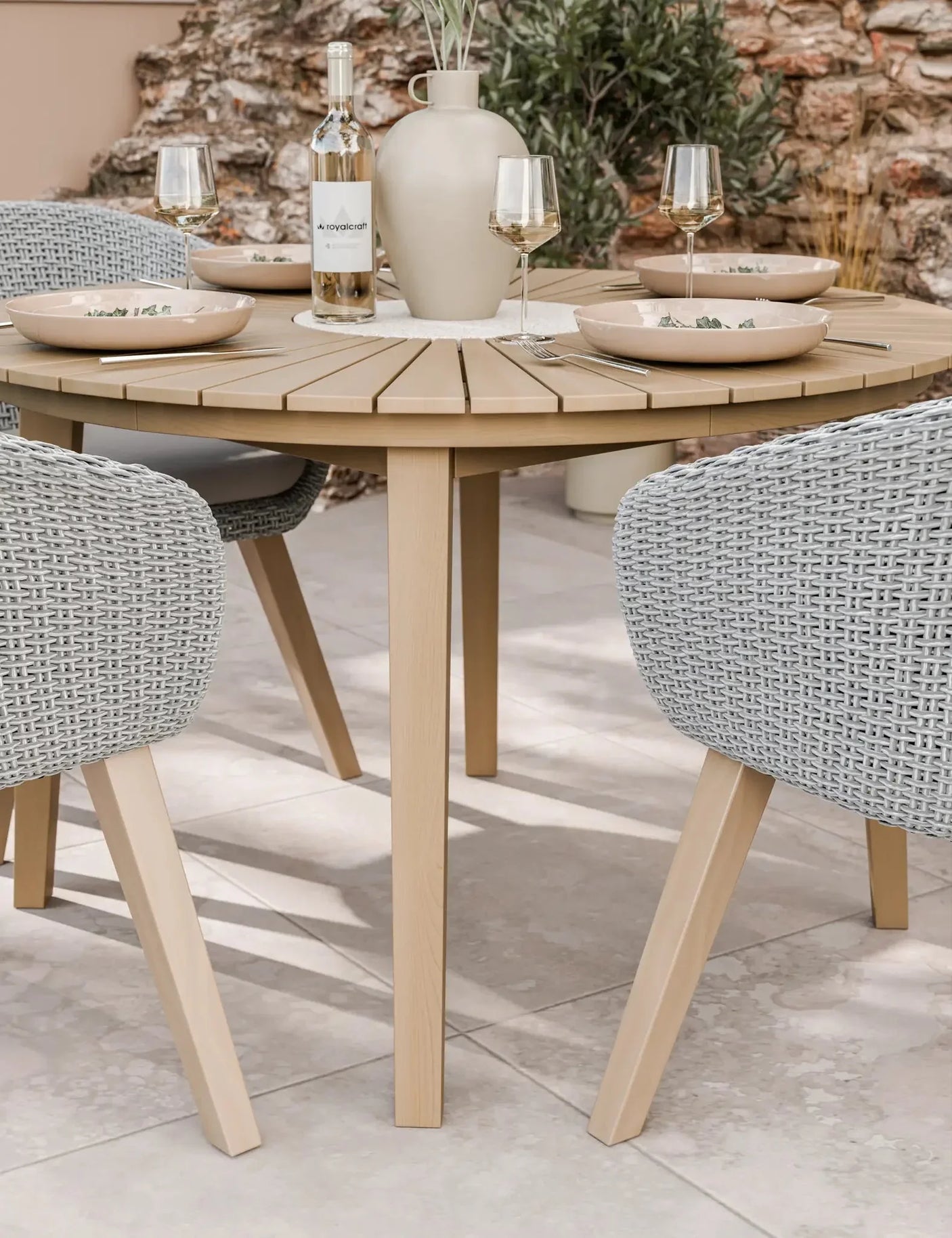 Round wooden outdoor dining table with wicker chairs, set for four with plates and glasses