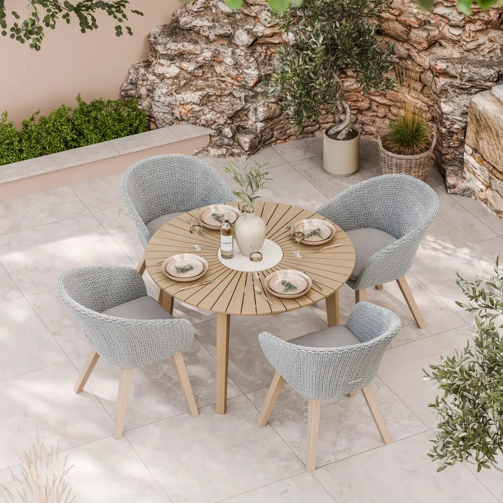 Round patio dining table with wood slats and four grey woven garden chairs on stone tiles