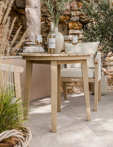 Light wood outdoor dining table with wine glasses and vase, patio stone backdrop