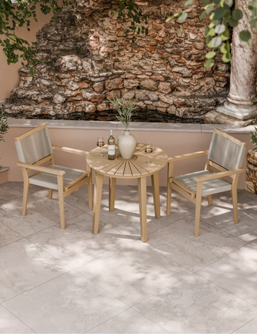 Outdoor bistro set with wooden table and two rope chairs on stone patio by a rock wall