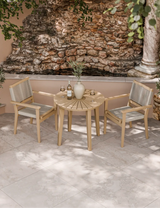Outdoor bistro set with wooden table and two rope chairs on stone patio by a rock wall