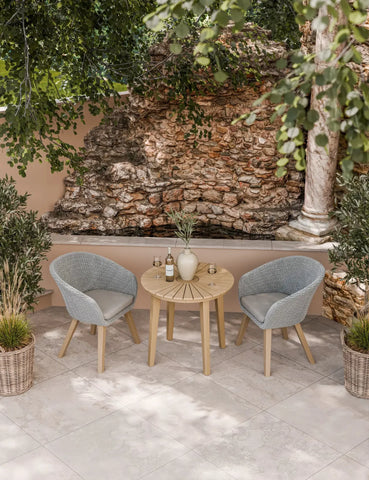 Outdoor bistro set with round wooden table and two grey rattan chairs on patio tiles