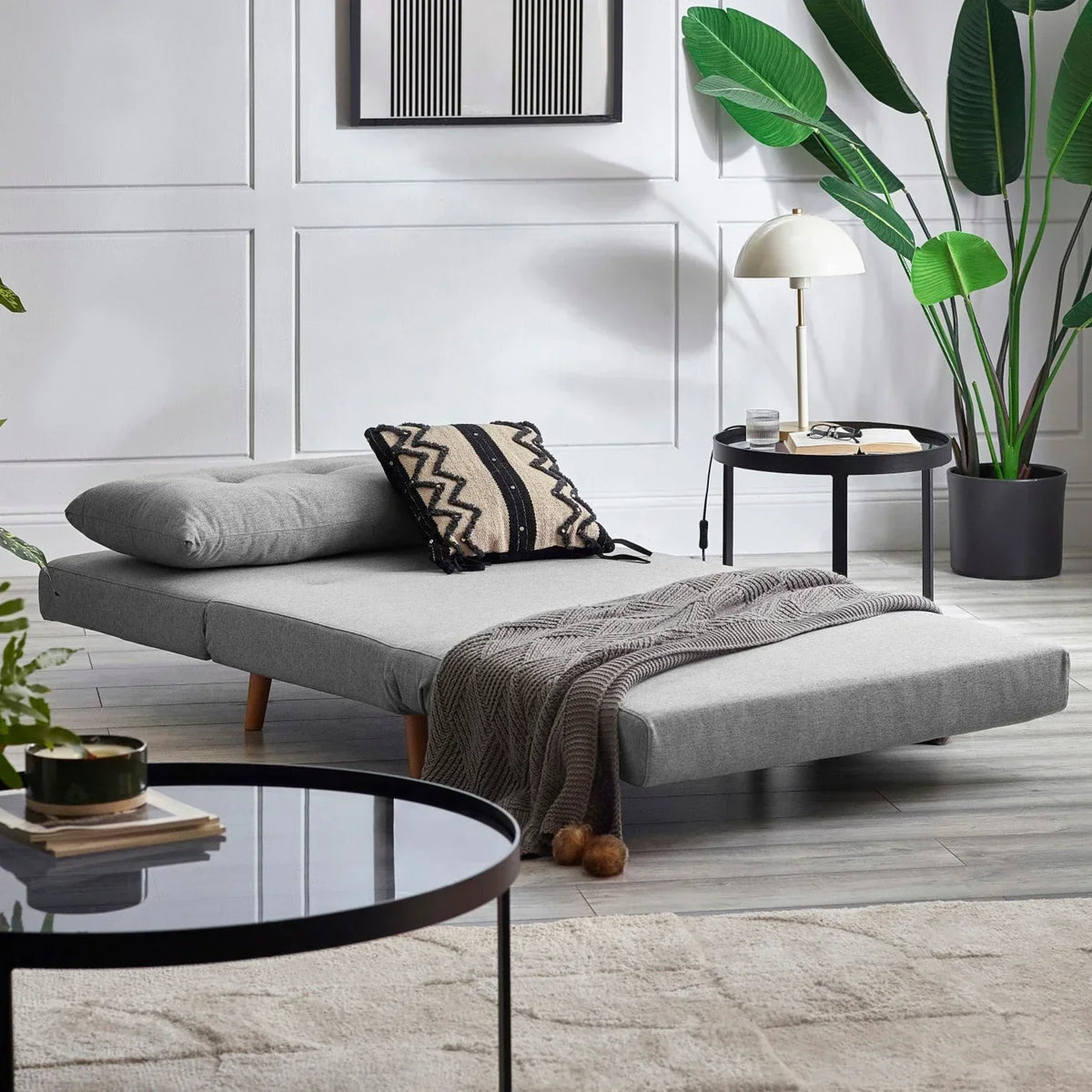 Grey single sofabed with cushion and throw in modern living room with plants and round tables