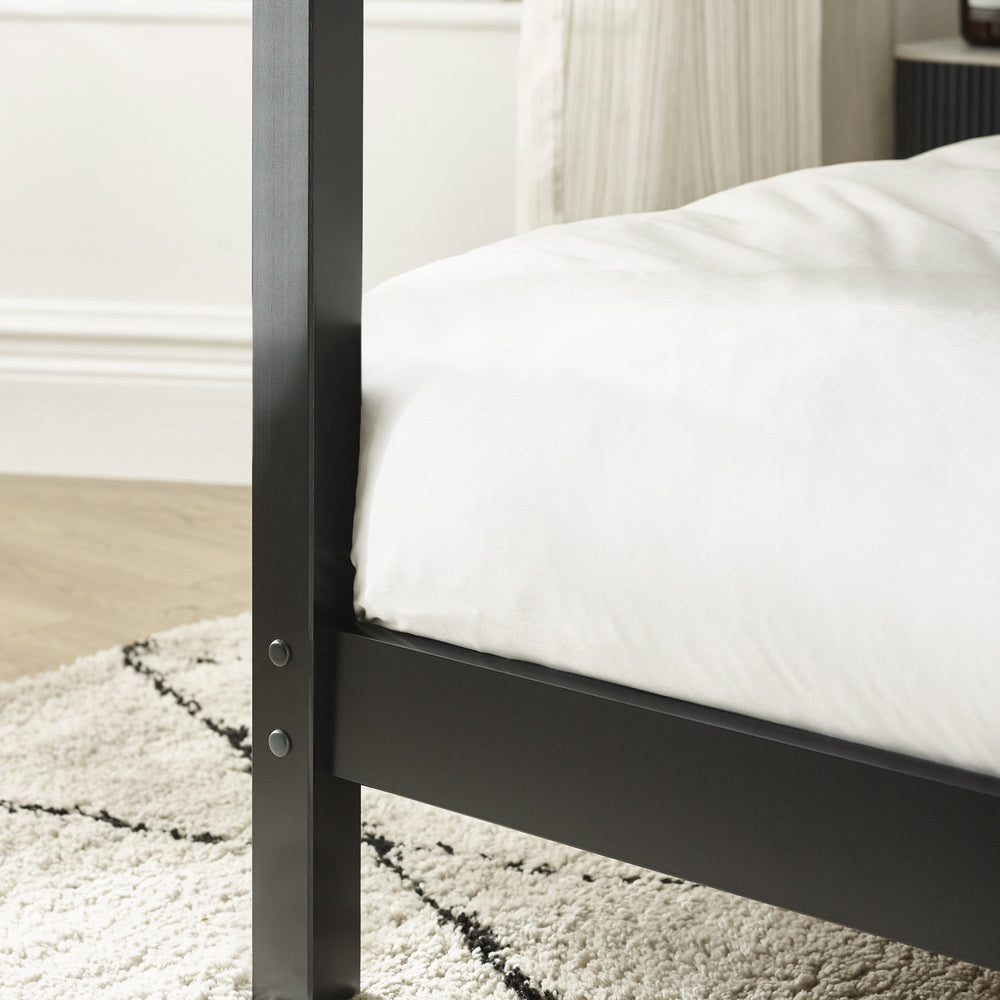 Close-up of black canopy bed post with metal bolts, light wood floor, and textured rug in background