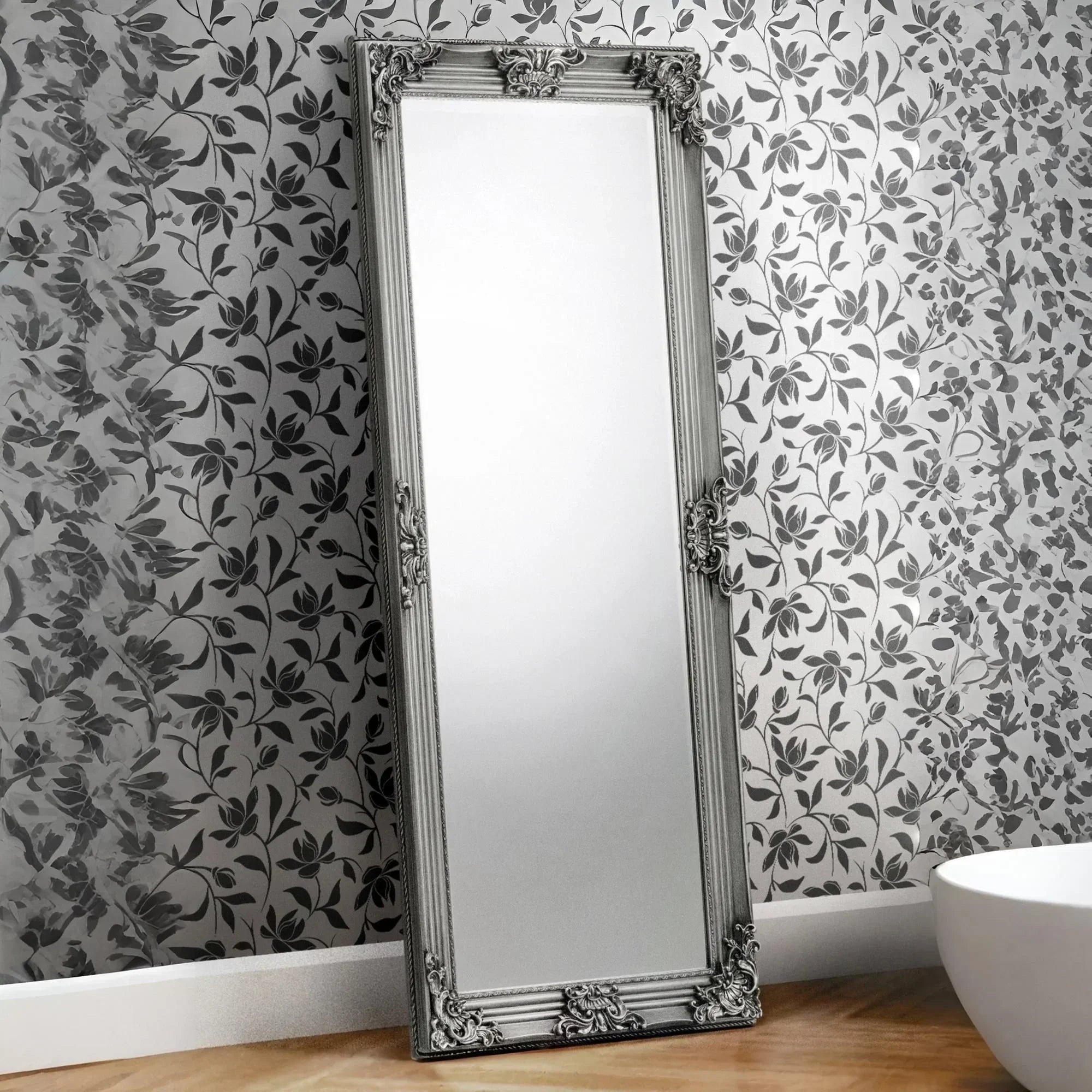Rococo full length mirror with ornate silver frame leaning against floral wallpapered wall