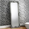 Rococo full length mirror with ornate silver frame leaning against floral wallpapered wall
