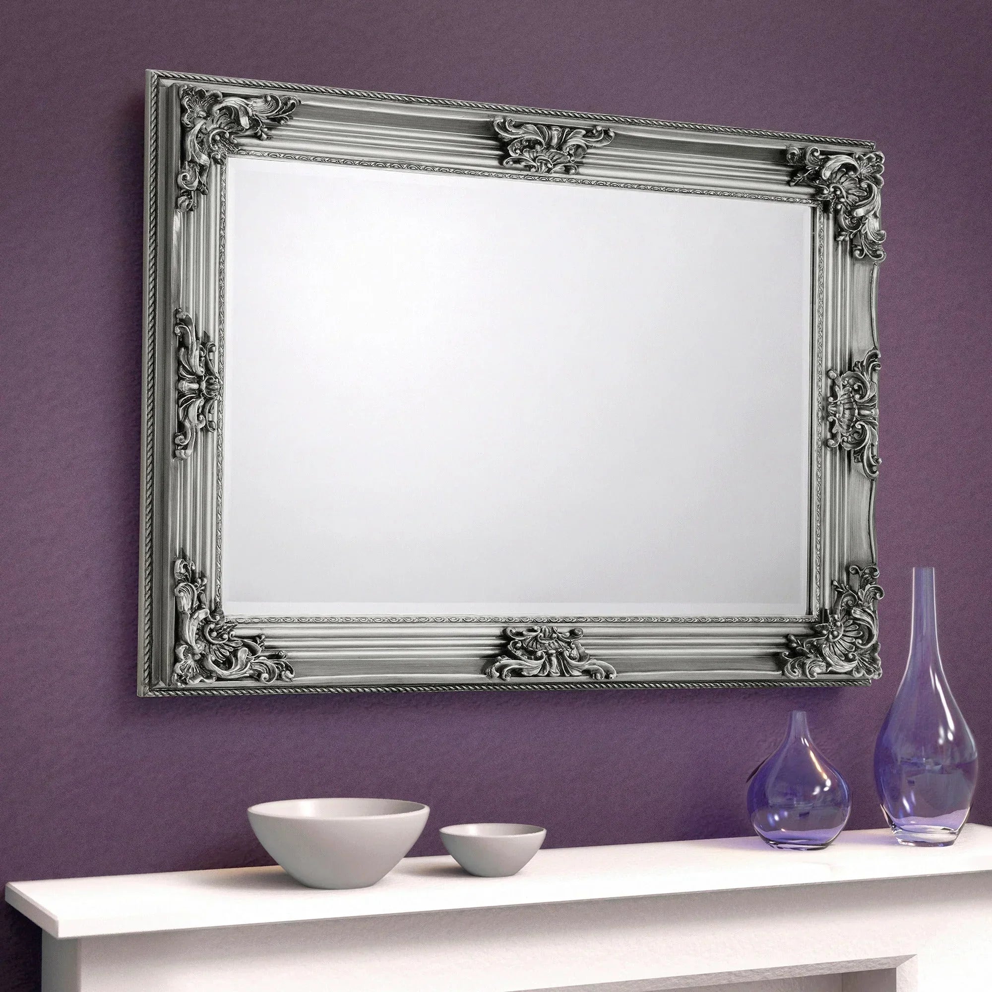 Rococo wall mirror with ornate silver frame above white mantel against purple wall
